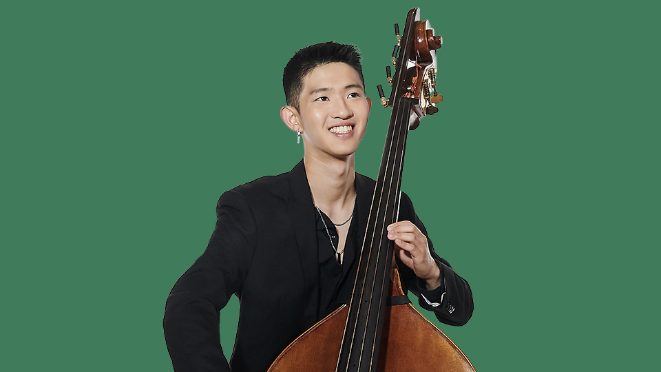 A man wearing a black shirt, holding a cello, smiling at the camera, with a dark green background.